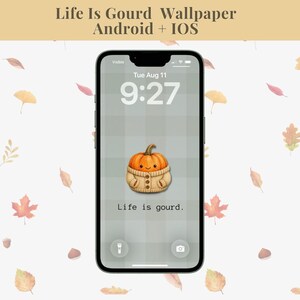 Op de afbeelding: Een smartphone met een digitale achtergrond genaamd Life Is Gourd Wallpaper Android + IOS. Het scherm toont een grijze ruitjesachtergrond met een centrale illustratie van een oranje pompoen in een beige gebreide trui en de tekst Life is gourd.