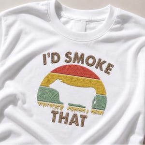 Può includere: Una t-shirt bianca con un ricamo raffigurante un tramonto a strisce rosse, gialle e verdi con la sagoma bianca di una mucca al centro. Il testo "I'D SMOKE THAT" è ricamato in marrone sopra e sotto la grafica del tramonto.