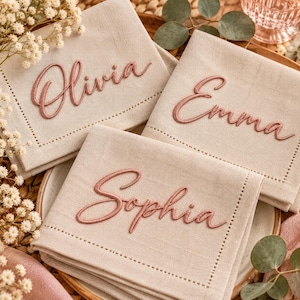 May include: Three beige linen napkins featuring embroidered names in a dusty rose script font. Each napkin displays a single name: Olivia, Emma, or Sophia, stitched in a cursive style with a decorative hemstitch border detail.