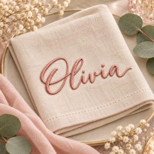 May include: A folded beige linen napkin featuring the name Olivia embroidered in a dusty rose script font. The napkin has a visible woven texture and a stitched border, presenting a clean and simple design for table settings.