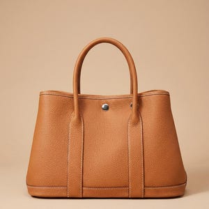 May include: A tan leather tote bag with a pebbled texture, featuring two vertical straps, a central silver-tone snap button closure, and two rounded top handles. The word ROVELLE is printed in the bottom right corner of the image.