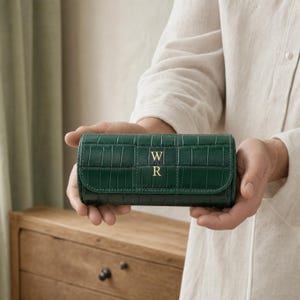 May include: A cylindrical dark green jewelry case with a crocodile-embossed texture. The front flap features the gold-colored letters W and R stacked vertically in the center. The case has a structured, rectangular shape with rounded edges.