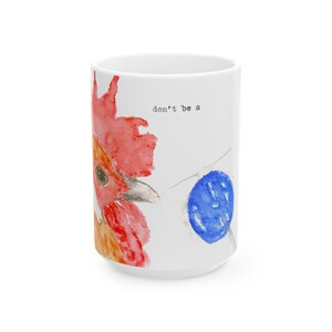 May include: A white ceramic mug featuring a watercolor illustration of a rooster head with a red comb and wattle. To the right of the rooster is a solid blue circle. The text "don't be a" is printed in small black typewriter-style font above the rooster.