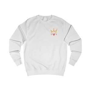 May include: A white long-sleeve sweatshirt featuring a small graphic on the upper left chest. The graphic consists of a gold-colored crown positioned directly above a small pink heart, with three short black lines radiating from each side.
