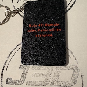 Könnte beinhalten: Ein rechteckiger schwarzer Schlüsselanhänger mit strukturierter Oberfläche und dem rot aufgedruckten Text Rule 47: Remain calm. Panic will be assigned. Der Anhänger ist mit einer kurzen silbernen Metallkette an einem silbernen Schlüsselring befestigt.