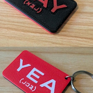 May include: A rectangular keyring with a metal ring. One side is black with red embossed text reading NAY and phonetic symbols. The other side is red with white embossed text reading YEA and phonetic symbols. The surface has a textured finish.