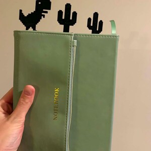 May include: A sage green notebook with a vertical zippered pocket on the front cover and the word NOTEBOOK printed in gold. Three black pixel-art style bookmarks shaped like a dinosaur and two cacti are inserted into the top of the notebook.