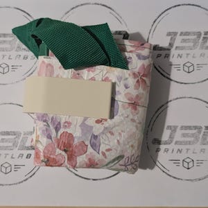 May include: A folded fabric item featuring a floral pattern with shades of pink, purple, and green. It includes a dark green woven strap and a solid off-white rectangular band wrapped horizontally around the center of the floral material.
