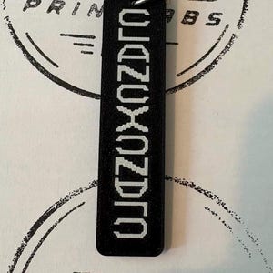Op de afbeelding: Een rechthoekige zwarte sleutelhanger met een getextureerd oppervlak met de witte tekst OJDZUXOZDPO verticaal gedrukt in een korrelig lettertype. De sleutelhanger is bevestigd aan een zilverkleurige metalen ring aan de bovenzijde.