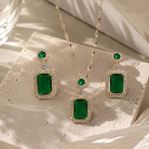 Op de afbeelding: Een goudkleurige sieradenset bestaande uit een ketting en twee oorbellen. Elk sieraad bevat een grote rechthoekige smaragdgroene steen omringd door een halo van kleine heldere kristallen, bekroond met een kleinere ronde groene steen.