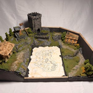 May include: A tabletop gaming diorama featuring a central parchment map, a grey stone tower, a small cottage with a thatched roof, and a wooden building. The landscape includes green mossy terrain, rocky cliffs, and miniature trees enclosed by a wooden frame.