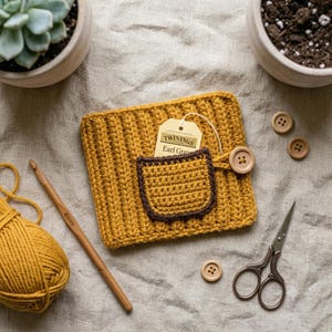 May include: A mustard yellow crocheted mug cozy with a ribbed texture and a small dark brown pocket. A Twinings Earl Grey tea tag is tucked into the pocket, which is secured by a wooden button. A wooden 4mm crochet hook, yarn, and scissors are nearby.