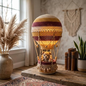 Op de afbeelding: Een gehaakte heteluchtballonlamp met horizontale strepen in crème, geel en bordeauxrood. De ballon is van binnenuit verlicht en bevat een kleine gehaakte teddybeer in de mand, die is versierd met houten kralen en kwastjes.