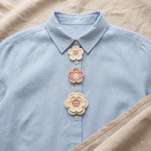 May include: A light blue button-down shirt featuring three crocheted flower embellishments attached to the front placket. The flowers are arranged vertically, alternating between cream and dusty rose colors with layered petals and centers.