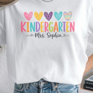 Puede incluir: Una camiseta blanca con un gráfico colorido que muestra cinco corazones estampados sobre la palabra KINDERGARTEN en letras multicolores. Debajo de la palabra, el texto Mrs. Sophia está escrito en cursiva negra con pequeños corazones a los lados.