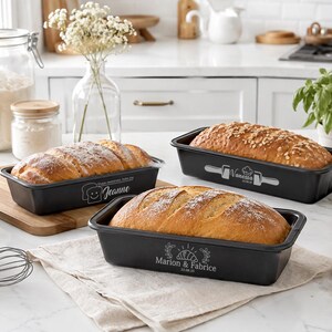 Pode incluir: Três formas retangulares de metal preto para pão contendo pão assado. As formas apresentam gravações brancas personalizadas: uma com um ícone de torrada e o nome Jeanne, uma com um rolo de massa e o nome Vanessa, e uma com um croissant e os nomes Marion & Fabrice.