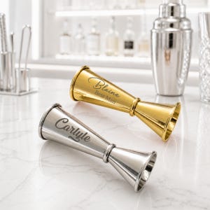 Pode incluir: Dois medidores de cocktail de dupla face, um dourado e outro prateado, ambos com acabamento metálico polido. O medidor dourado está gravado com o texto Blaine Best Man, enquanto o prateado apresenta o nome Carlyle em letra cursiva.
