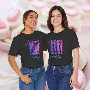 Puede incluir: Una camiseta de manga corta gris oscuro con el texto Team BRIDE en letras azules y rosas apiladas, con el nombre Ashley impreso en una fuente cursiva de color verde azulado debajo. La camiseta se combina con vaqueros azules.
