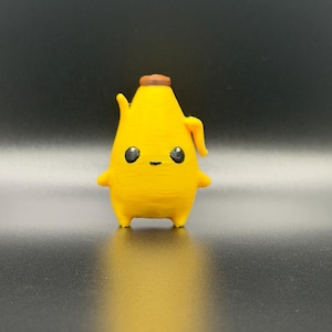 May include: A small, yellow, banana-shaped figurine with a brown stem at the top. The figure has a smooth, rounded body, two small arms extending from the sides, and two short legs. It features a printed face with two large, black, glossy eyes and a small, thin, horizontal line for a mouth.