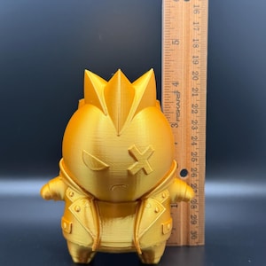 May include: A metallic gold 3D-printed figurine of a character with a mohawk, wearing a jacket, and featuring an X-shaped eye. The figurine is positioned next to a wooden ruler, showing a height of approximately 3.5 inches from base to top.