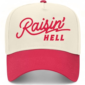 Op de afbeelding: Een crèmekleurige baseballpet met een felrode klep en een rode afbeelding op de voorkant. De tekst "Raisin' HELL" is in een gestileerd handschrift en blokletters in het midden van de kroon gedrukt, voorzien van een kleine rode knoop.