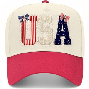 May include: A cream-coloured baseball cap with a bright pink brim. The front features the letters USA in a decorative style: the U has red and white stripes with a blue star bow, the S is textured cream, and the A is blue with white stars and a red and white striped bow.