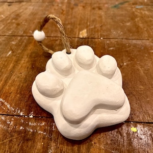 May include: A white clay paw print ornament featuring a textured surface shaped like an animal paw. The ornament includes a natural jute twine loop for hanging, accented with a single small white wooden bead attached to the top of the clay piece.