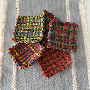 May include: A set of four square woven coasters featuring a multicolored grid pattern in shades of blue, orange, green, yellow, and red. Each coaster has a thick, braided border and a textured, interlocking weave construction made of fabric strips.