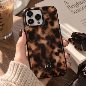 May include: A smartphone case featuring a brown and black tortoiseshell pattern with the gold-colored initials HT printed at the bottom. The case has a cutout for a triple-lens camera system and is designed to fit a rectangular mobile device.