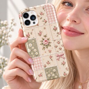 May include: A smartphone case featuring a patchwork design of cream, sage green, and soft pink squares. The pattern includes illustrations of small roses, daisies, and a delicate pink bow. The case has a cutout for a triple-lens camera system.