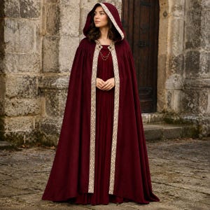 May include: A floor-length, deep burgundy hooded cloak featuring a wide, ornate cream-colored trim along the front edges and hood. The garment includes a decorative metal clasp at the neckline with two thin, dangling gold-toned chains.