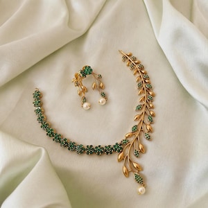 May include: A gold-toned necklace and earring set featuring a floral and leaf motif. The necklace has a chain of small green stone flowers on one side and a large, sweeping gold leaf branch on the other, accented with a single white pearl.
