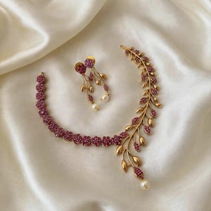 May include: A gold-toned necklace and earring set featuring a floral design with small pink stones. The necklace has a curved branch-like section with gold leaves and pink stones, ending in a white pearl. The earrings feature similar floral motifs.
