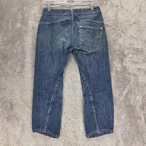 May include: A pair of blue denim jeans featuring a faded wash, a single back pocket on the right side with a small red tab, and a visible seam construction running down the legs. The jeans are displayed from the back view with a metal hanger.