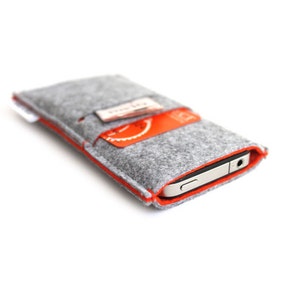 Könnte beinhalten: Ein graues Filz-Handy-Etui mit orangefarbenem Stepprand. Das Etui hat einen Steckplatz für eine Kreditkarte und ein Telefon ist im Inneren sichtbar.