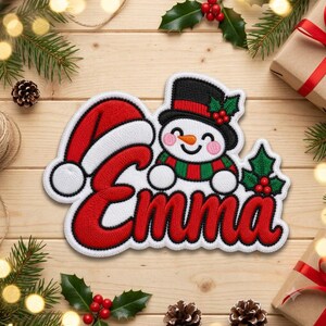Puede incluir: Un parche bordado con el nombre Emma en letra roja y borde blanco. Un muñeco de nieve con sombrero de copa negro, acebo y bufanda roja y verde se sitúa sobre el nombre. Un gorro de Papá Noel rojo está unido a la letra E.