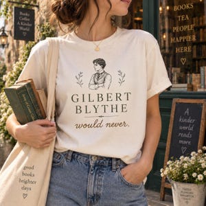 May include: A cream-colored short-sleeve t-shirt featuring a black line-art illustration of a man in a vest, with the text "GILBERT BLYTHE would never." printed in black and brown. The shirt is paired with blue denim jeans and a canvas tote bag.