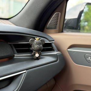 May include: A black bear-shaped car air freshener clip covered in small dark crystals. The bear wears a small gold-coloured crown on its head and a circular pendant featuring the letter J hanging from a chain around its neck.