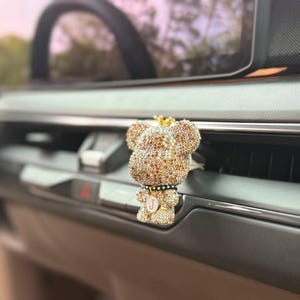 Puede incluir: Un ambientador de coche con clip de ventilación en forma de oso, cubierto de brillantes pedrería dorada y bronce. El oso presenta una pequeña corona dorada en la cabeza, un collar negro con un colgante circular y un centro blanco con la letra J.