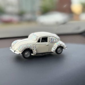 May include: A miniature off-white toy car model featuring a classic rounded silhouette, adorned with numerous small, reflective, iridescent crystals covering the front fenders, the hood area, and the rear wheel arches, with black wheels.