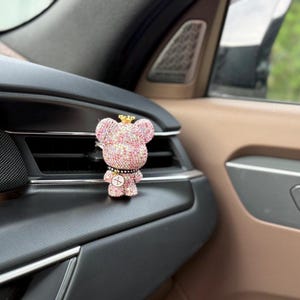 May include: A small bear-shaped car air freshener clip covered in sparkling pink and white rhinestones. The bear features a gold-toned crown on its head, a black collar with a small circular charm, and is attached to a vehicle air vent.