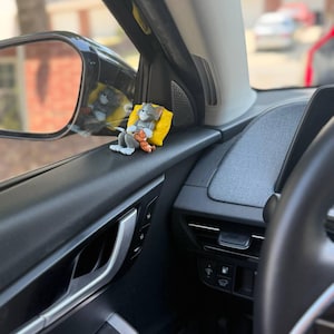 May include: A small figurine of the cartoon character Tom the cat resting against a bright yellow pillow. The grey cat is depicted in a relaxed, reclining position with a small brown teddy bear tucked under one arm, placed on a car window ledge.
