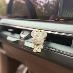 May include: A car air freshener shaped like a bear figure covered in small, sparkling clear crystals. The bear wears a small gold crown on its head and a black collar around its neck with a circular pendant featuring the letter Q in the centre.