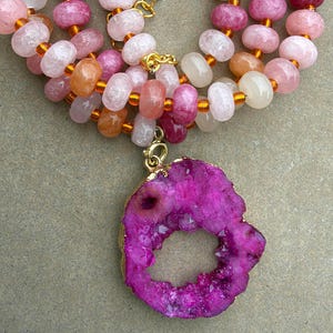 May include: A necklace featuring a large, irregular, vibrant magenta geode pendant with a gold-toned edge. The strand consists of round, translucent beads in shades of pink, peach, and white, interspersed with small, transparent amber-colored spacer beads.
