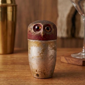May include: A decorative cocktail shaker with an owl design. The top is a dark red color with large, amber-colored eyes. The bottom half is a metallic silver with gold accents. The shaker is made of metal and sits on a wooden surface.