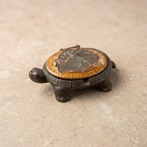May include: A cast iron turtle-shaped ashtray with a hinged shell lid. The lid features a worn, yellow and brown patterned border with the text AKRON OHIO visible at the bottom. The metal body is dark brown with a textured, weathered finish.