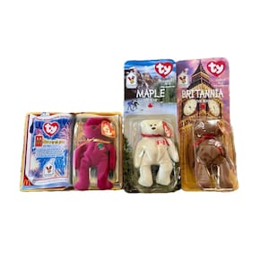 May include: A collection of Ty Beanie Babies, including a magenta bear, a white bear with a Canadian flag, and a brown bear with a red bow. The packaging features the Ty heart logo and country-themed designs.