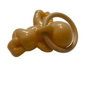 May include: A light peach-colored plastic baby figurine in a crawling position, featuring a smooth, glossy finish. The figurine is depicted holding a circular ring in its hands, with a rounded head and defined limbs, crafted from a single molded piece.
