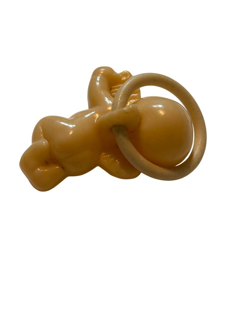 May include: A light peach-colored plastic baby figurine in a crawling position, featuring a smooth, glossy finish. The figurine is depicted holding a circular ring in its hands, with a rounded head and defined limbs, crafted from a single molded piece.