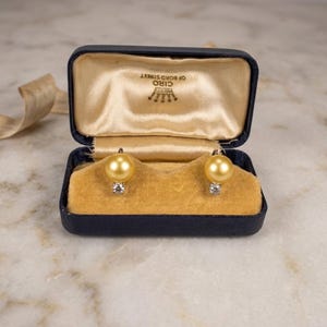May include: A pair of gold-toned pearl earrings with a single clear stone accent, presented in a navy blue rectangular jewelry box. The interior lid features the text CIRO OF BOND STREET with a crown logo, lined with cream satin and gold velvet.